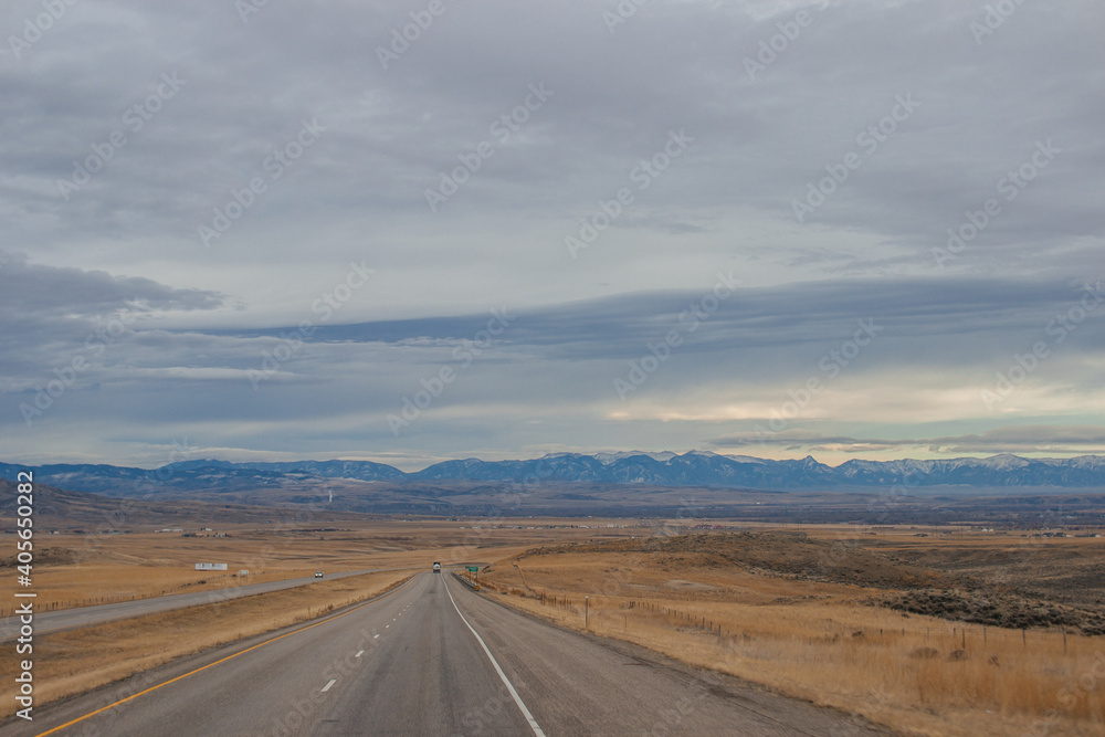 Asphalt road among steppes with yellow dry grass, blue mountains on the horizon, beautiful blue-gray autumn sky. Montana, USA, 11-23-2019