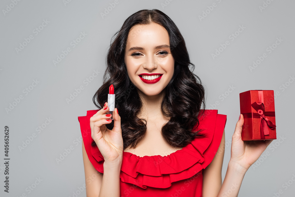 cheerful young woman holding red lipstick and wrapped present isolated on grey cheerful young woman holding red lipstick and wrapped present isolated on grey