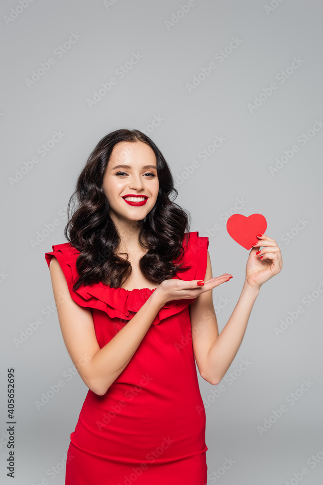 happy young woman pointing with hand at red paper heart isolated on grey happy young woman pointing with hand at red paper heart isolated on grey