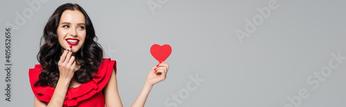 cheerful young woman holding red paper heart and lipstick isolated on grey, banner