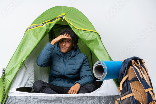 Young african american man inside a camping green tent looking far away with hand to look something