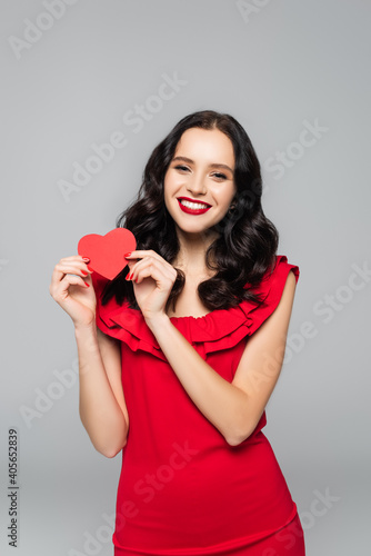 cheerful young woman in dress holding red paper heart isolated on grey
