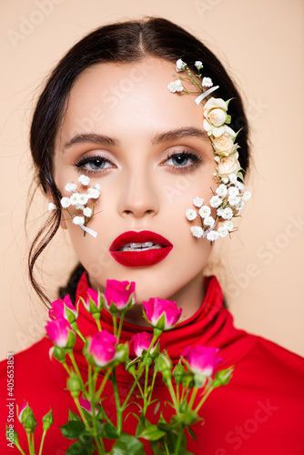 young woman with red lips and flowers on face looking at camera isolated on pink