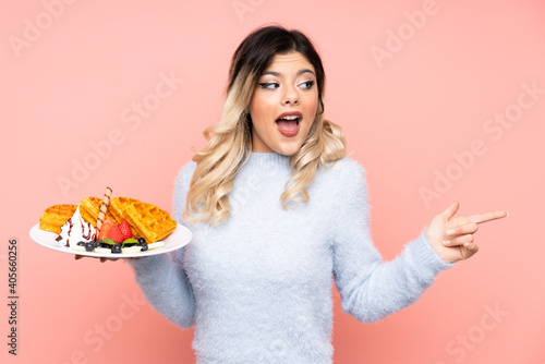 Teenager girl holding waffles on isolated pink background surprised and pointing finger to the side