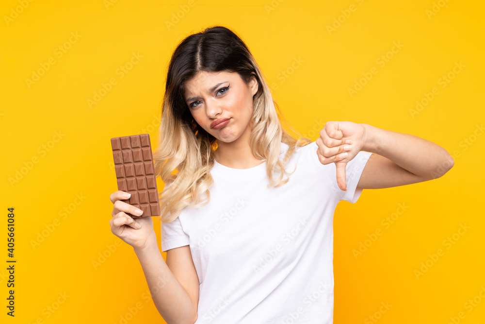 Teenager girl isolated on yellow background taking a chocolate tablet making bad signal Teenager girl isolated on yellow background taking a chocolate tablet making bad signal