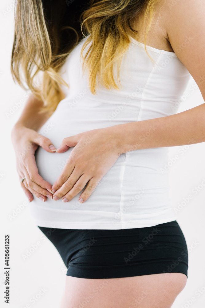 Pregnant woman in cotton underwear standing indoors with hands resting on belly in heart shape Pregnant woman in cotton underwear standing indoors with hands resting on belly in heart shape