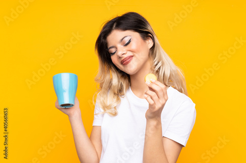 Teenager girl isolated on yellow background holding colorful French macarons and a cup of milk