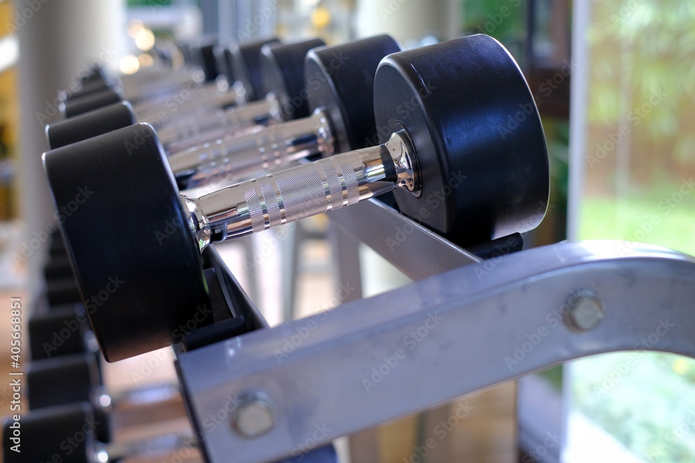 A close-up picture of steel dumbbells with black rubber protection lining up in rows on a metal rack in a gym by a glass window. A close-up picture of steel dumbbells with black rubber protection lining up in rows on a metal rack in a gym by a glass window.