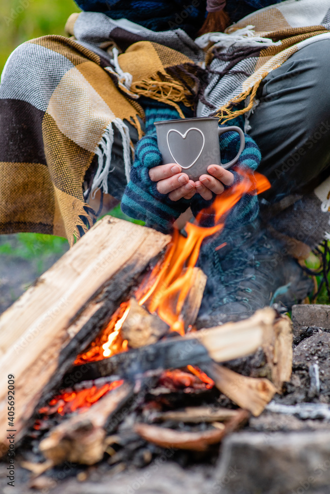 Obraz premium tourist wrapped a plaid holds cup of the hot tea near a bonfire