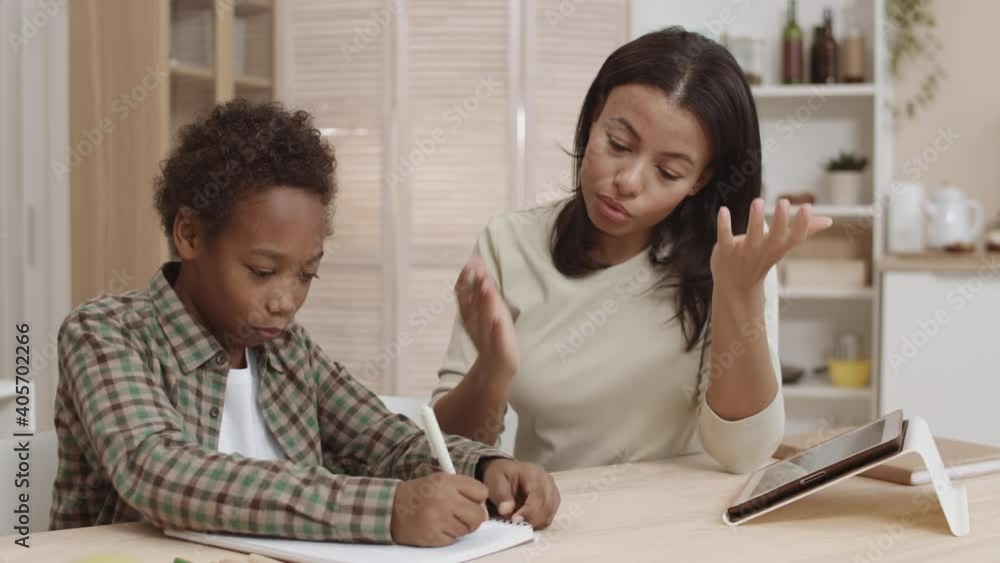 Medium close-up of irritated Mixed-Race mother sitting by desk at home next to guilty African son, scolding him for making mistakes doing homework, then standing up and leaving Medium close-up of irritated Mixed-Race mother sitting by desk at home next to guilty African son, scolding him for making mistakes doing homework, then standing up and leaving