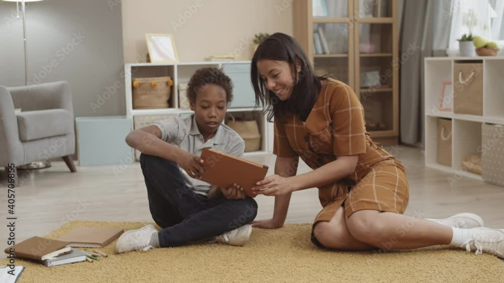 Full shot of Mixed-Race mother smiling, looking at tablet computer her African school-aged son showing her. Diverse family members sitting on floor at home, having fun together Full shot of Mixed-Race mother smiling, looking at tablet computer her African school-aged son showing her. Diverse family members sitting on floor at home, having fun together