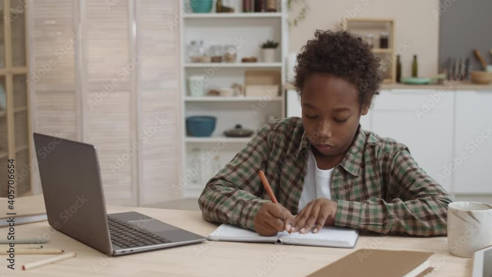Chest-up of African ten-year-old male student sitting by desk at home, writing in notebook, looking at laptop computer, then getting tired of doing difficult homework, putting head down on hands Chest-up of African ten-year-old male student sitting by desk at home, writing in notebook, looking at laptop computer, then getting tired of doing difficult homework, putting head down on hands