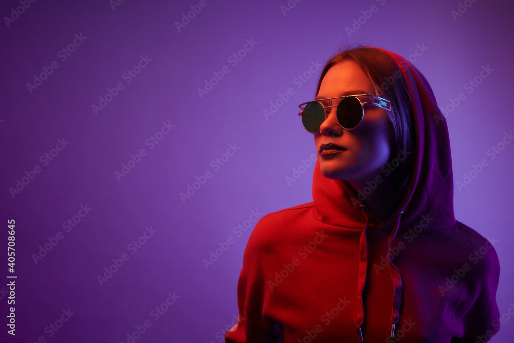 Young model posing in a studio in a trendy neon light. Woman in round sunglasses and hoodie with colorful light effects. Young model posing in a studio in a trendy neon light. Woman in round sunglasses and hoodie with colorful light effects.