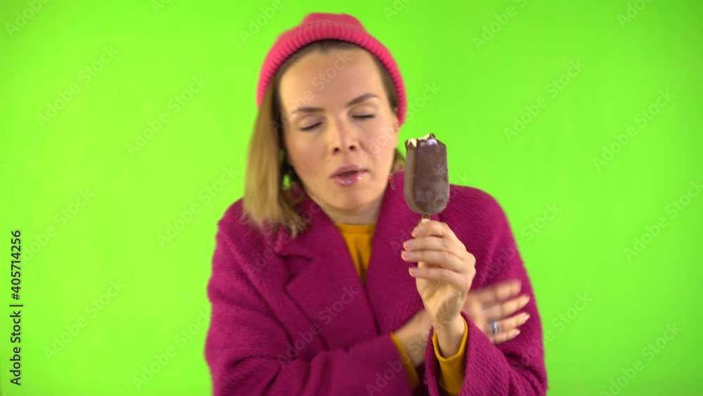 Woman Bites Ice Cream and Frozen, She Shakes from the Cold. Woman Dressed in Knitted Hat, Red Coat on Green Background in Studio. Woman Bites Ice Cream and Frozen, She Shakes from the Cold. Woman Dressed in Knitted Hat, Red Coat on Green Background in Studio.