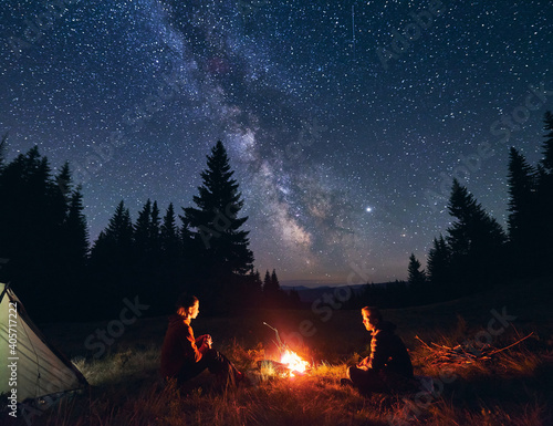 Man and woman communicate by fire against background of starry sky with bright Milky way. Night camping. Silhouettes of big fir trees against backdrop of mountain valley under an incredible sky