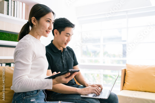 Asian woman using a tablet and looking with confidence, sitting by the man working on the laptop. Casual businesspeople working together at home. The successful woman leading in business.