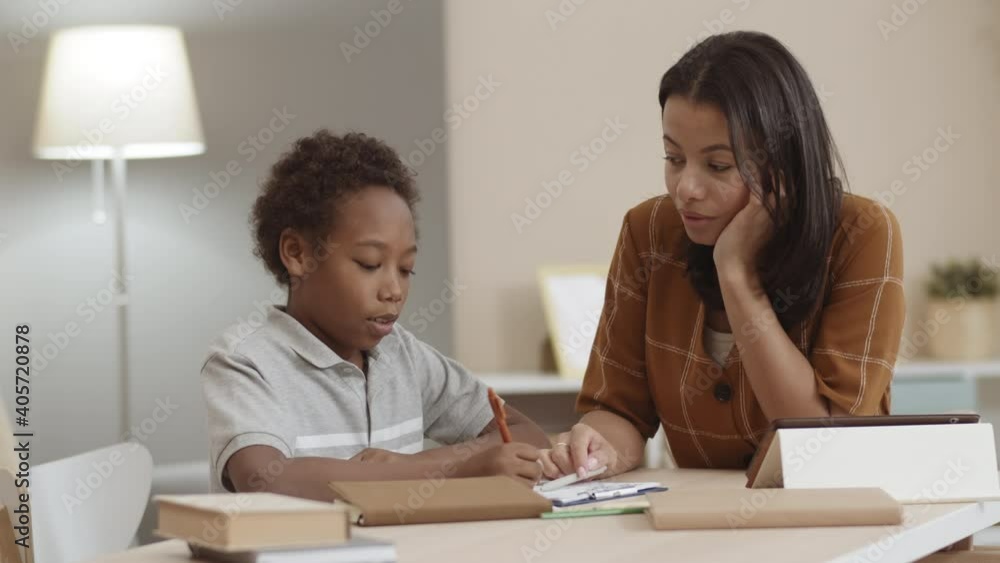 Medium close-up slow motion of young Mixed-Race mother helping African school boy studying at home. Diverse family members sitting by desk, talking Medium close-up slow motion of young Mixed-Race mother helping African school boy studying at home. Diverse family members sitting by desk, talking