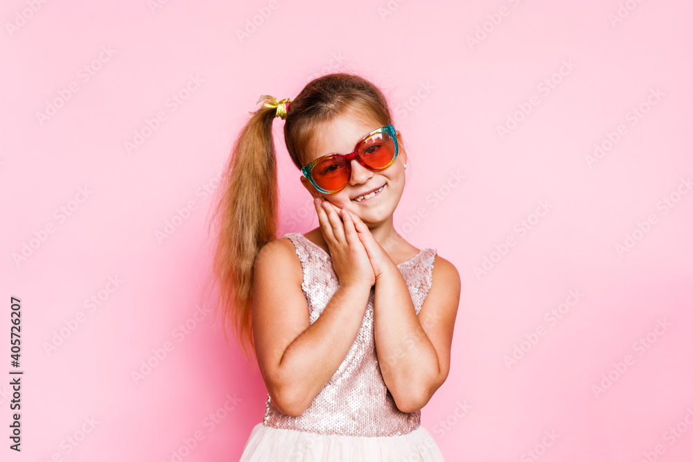 Girl with long hair in festive paper hat and dressy dress on pink background tossing gold confetti Girl with long hair in festive paper hat and dressy dress on pink background tossing gold confetti