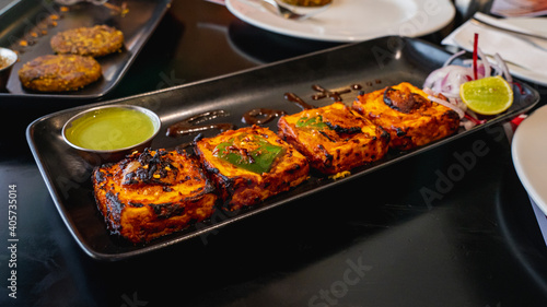 Selective focus of Paneer Tikka Kebab served with green chutney on a black serving tray. White plate, Potato Quinoa Patty on an appetizer tray blurred in the background.