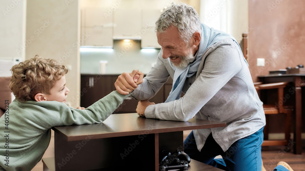 Who is stronger. Happy grandfather and little grandson sitting opposite each other and playing arm wrestling, having fun at home Who is stronger. Happy grandfather and little grandson sitting opposite each other and playing arm wrestling, having fun at home