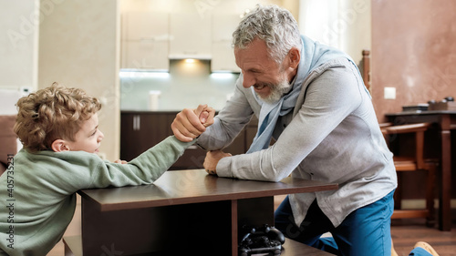 Who is stronger. Happy grandfather and little grandson sitting opposite each other and playing arm wrestling, having fun at home