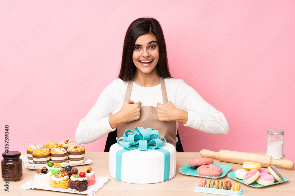 Pastry chef with a big cake in a table over isolated pink background with surprise facial expression Pastry chef with a big cake in a table over isolated pink background with surprise facial expression