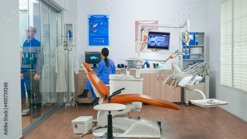 Nurse inviting next patient in stomatology room showing to lie on chair. Dentistry assistant sitting in consultation dental room with elderly woman while doctor speaking with old man in background. © DC Studio