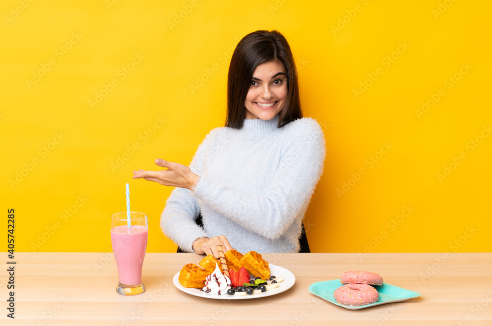 Young woman eating waffles and milkshake in a table over isolated yellow background extending hands to the side for inviting to come Young woman eating waffles and milkshake in a table over isolated yellow background extending hands to the side for inviting to come