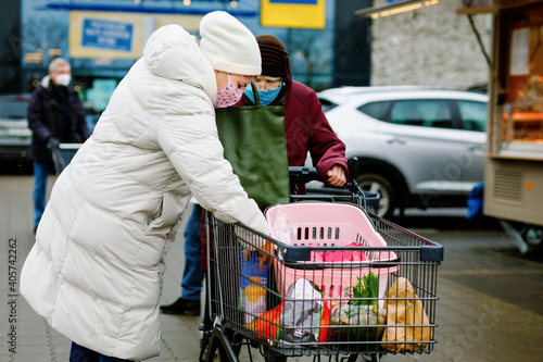 Senior woman and social worker with medical mask due pandemic coronavirus disease. Daughter or granddaughter help grandmother with shopping in supermarket, push cart trolley with foods, outdoors © Irina Schmidt