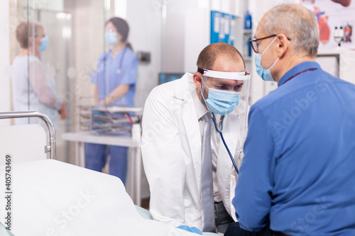 Cardiologist in hospital checking senior man heart illness using stethoscope in covid19. Medical practitioner wearing face mask consulting senior man in examination room during coronavirus.