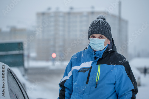Old man wearing knitted cap and protective face mask stands near car on roadside