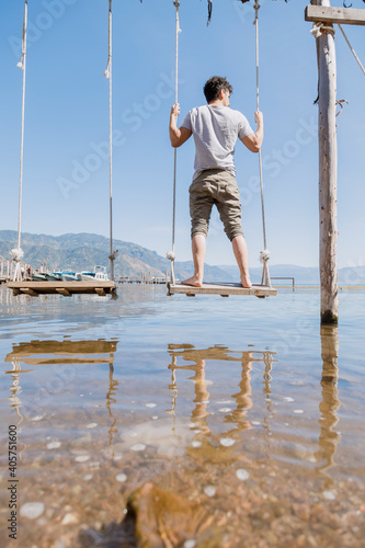 Fototapeta Naklejka Na Ścianę i Meble -  Young man standing on a swing in the middle of Lake Atitlan - Hispanic traveler enjoying Lake Atitlan in Guatemala - Lake landscape in the morning