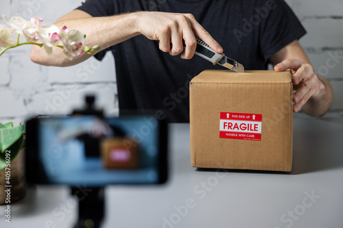 man recording unboxing video with mobile phone. cutting cardboard box with knife. social media marketing photo