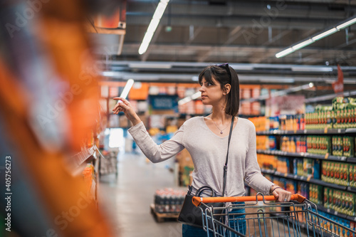 Sale, shopping, consumerism and people concept. Woman in supermarket with shopping trolley choosing products and looking aside. Female chooses products on the background of shelvesin a supermarket. 