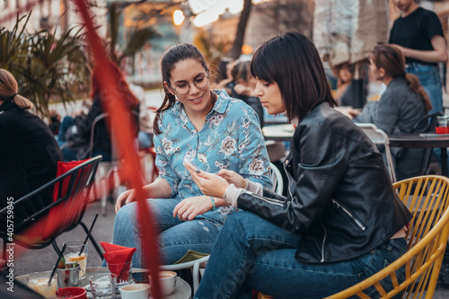 Two beautiful women in cafe outdoors looking at something on mobile phone. Two female using mobile phone application shopping online and enjoying coffee outside in city
