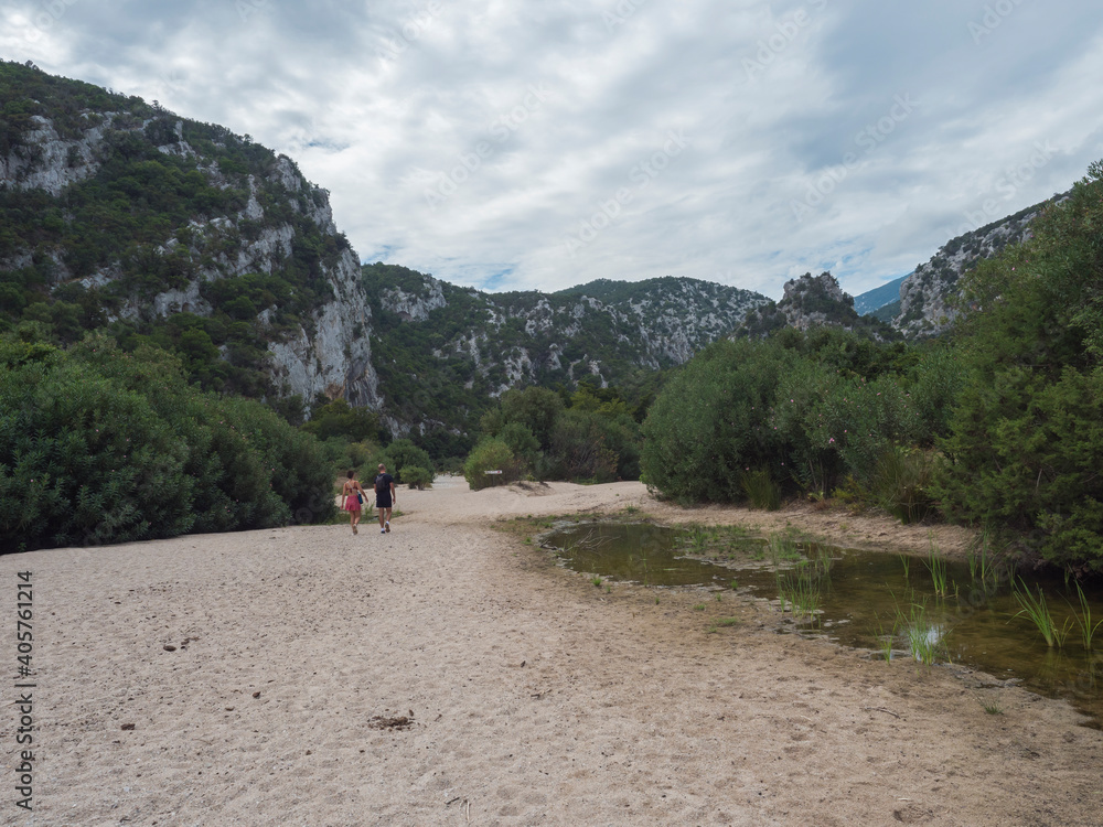 Man and woman couple arrivinag on sand path to Cala Luna beach at Gulf of Orosei with green bushes, trees and limestone rocks. Sardinia, Italy.