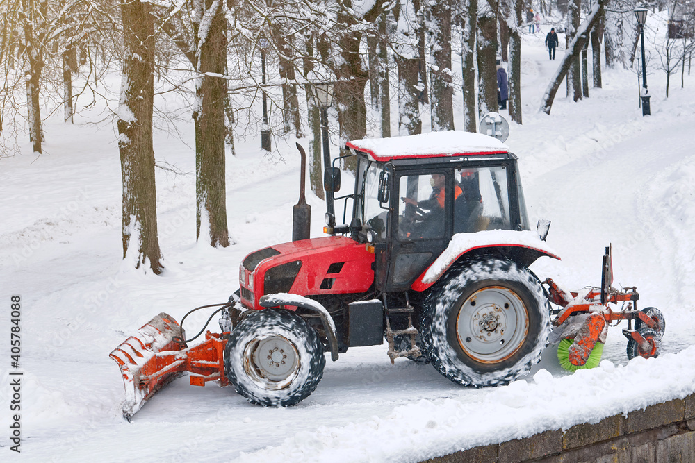 Snow removal tractor with scoop and rotating brush sweeping snow from footpath in park. Red tractor automated rotating brush removes snow from walkway in city park. Municipal road sweeping vehicle Snow removal tractor with scoop and rotating brush sweeping snow from footpath in park. Red tractor automated rotating brush removes snow from walkway in city park. Municipal road sweeping vehicle
