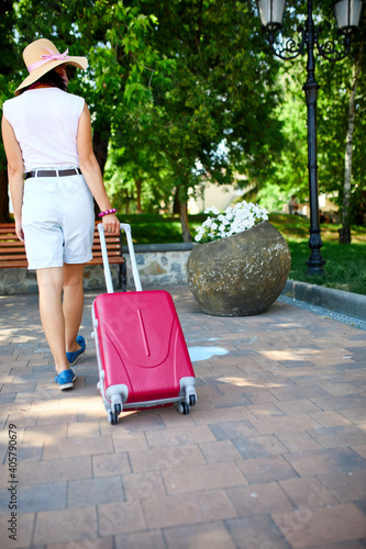 Woman in straw hat and pink protective mask, walks down the street with a suitcase