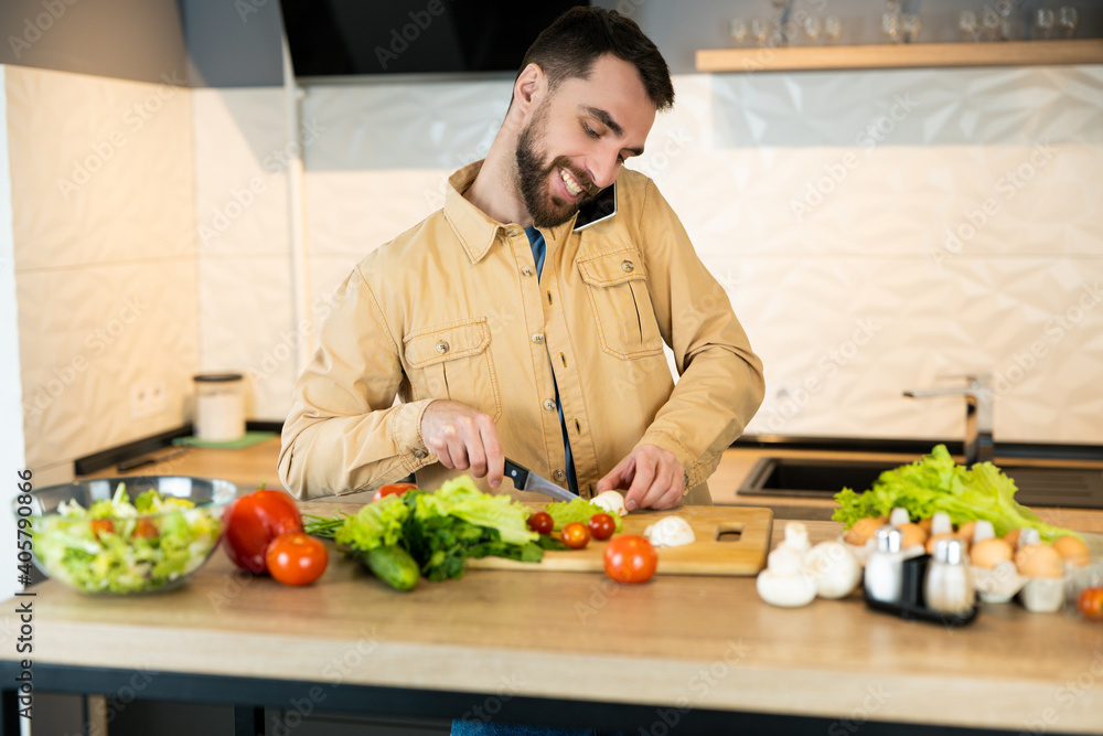 Fototapeta premium Young man is talking on phone while cooking salad in the kitchen. Vegan guy is cutting vegetables for the salad.