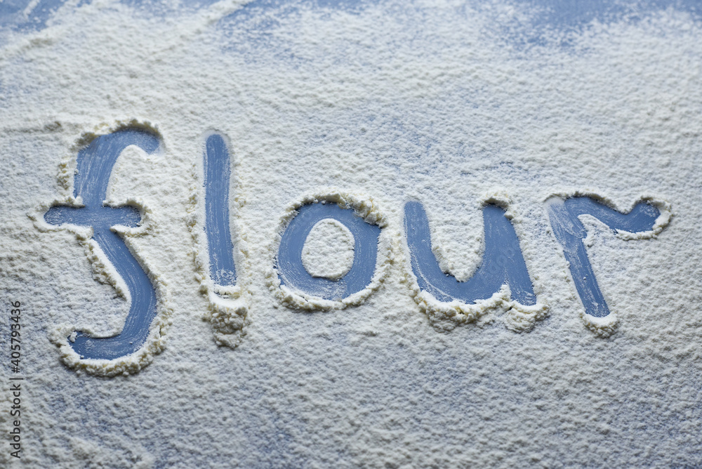 The word flour is written on scattered flour on a blue background. The word flour is written on scattered flour on a blue background.