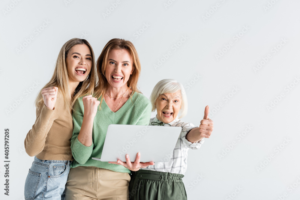 Obraz premium three generation of happy women looking at camera, showing thumb up and holding laptop isolated on white