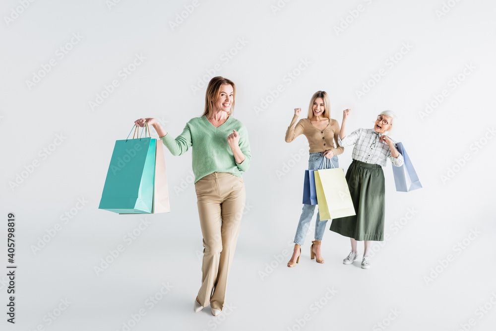 Naklejka premium full length of happy woman with shopping bags near daughter and senior mother on blurred white background