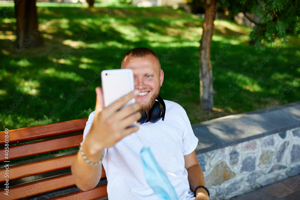 Man in medical mask sitting on the bench in the park take selfie by mobile phone. Man in medical mask sitting on the bench in the park take selfie by mobile phone.