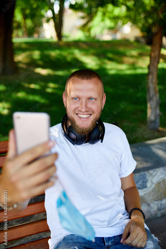 Man in medical mask sitting on the bench in the park take selfie by mobile phone. Man in medical mask sitting on the bench in the park take selfie by mobile phone.