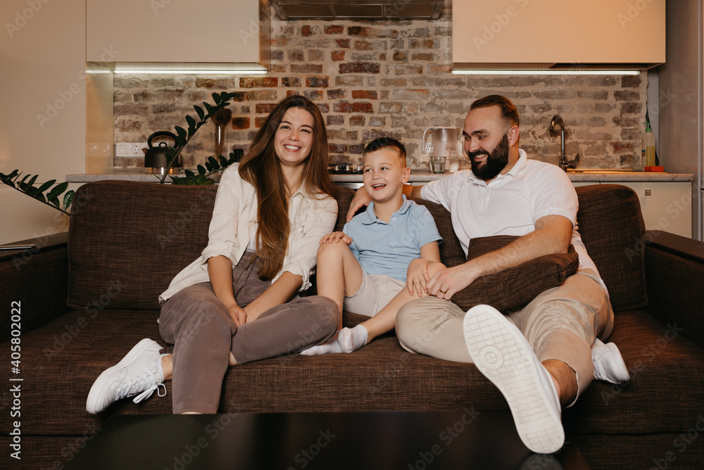 Dad, son, and mom are watching TV and laughing on the sofa in the apartment. The family is enjoying a happy evening at home. The father is staring at his boy and wife. Dad, son, and mom are watching TV and laughing on the sofa in the apartment. The family is enjoying a happy evening at home. The father is staring at his boy and wife.