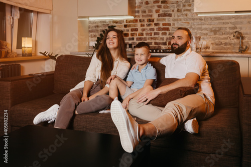 Dad with a beard, son, and young mom with long hair are watching with interest TV and smiling on the sofa in the apartment. The family is enjoying a happy evening at home.