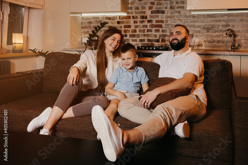 Dad with a beard, son, and young mom with long hair are watching TV and smiling on the sofa. The family is enjoying a happy evening at home. The mother is laughing