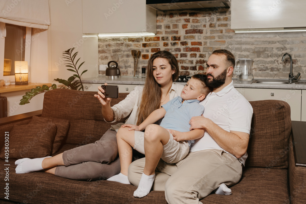 A dad, a son, and a young mother are watching a video on a smartphone on the sofa. A mom is demonstrating to her family news on the screen of the cellphone in the evening at home.