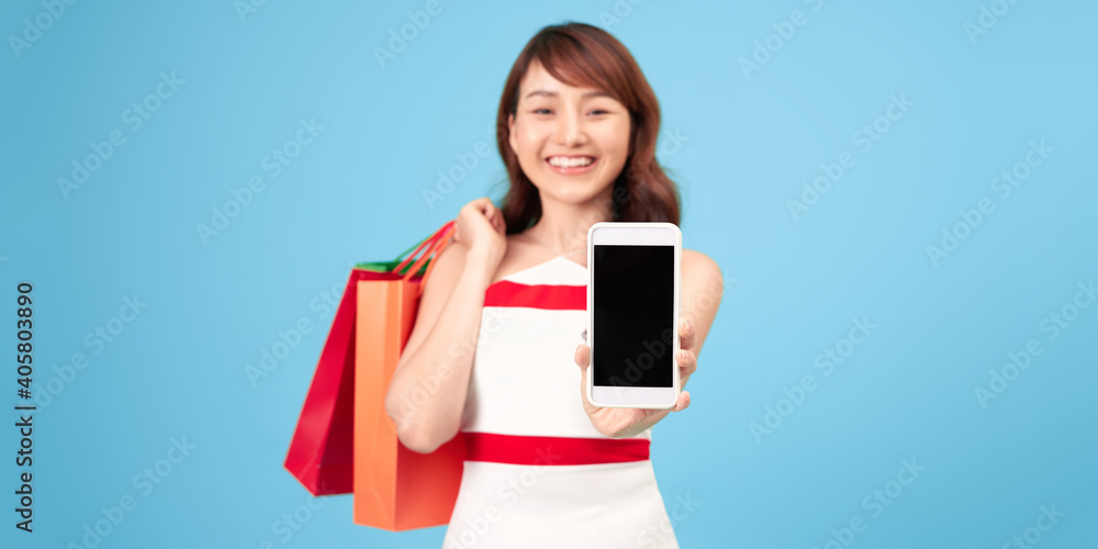 Happy lady with colored paper bags shows smartphone with blank screen and takes order isolated on blue background Happy lady with colored paper bags shows smartphone with blank screen and takes order isolated on blue background