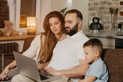 A father with a beard is talking about his achievements at work to his son and wife at home. A young family on the sofa in the evening. Dad is working remotely on a laptop between relatives